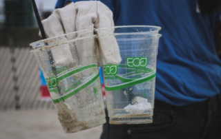 A gloved hand is holding up several clear plastic cups that have been printed with Eco in green on the side. They are covered in sand like they were picked up off a beach.